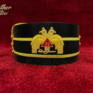 Puede incluir: Gorra negra con bordados y adornos dorados. La gorra presenta un emblema dorado con dos águilas y un triángulo rojo con el número 32. Borlas doradas cuelgan del lateral. La gorra se exhibe sobre una superficie roja.