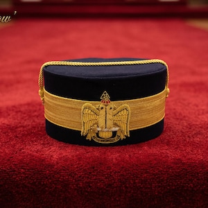 Puede incluir: Un sombrero azul marino con ribetes dorados y un emblema dorado de un águila. El sombrero cilíndrico está sobre una alfombra roja. El texto "Leather Stow" está en la esquina superior izquierda.