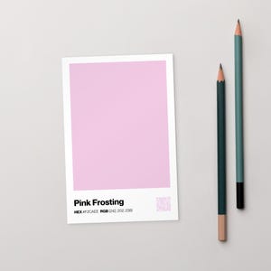 Può includere: Una scheda rettangolare bianca con un campione di colore rosa etichettato "Pink Frosting" e informazioni sul codice colore. Due matite, una verde acqua e una verde scuro, sono posizionate a destra della scheda.