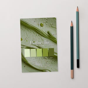 May include: A "Pistachio" colour palette card with a green, ice cream-like background and a colour swatch. The card displays a range of green hues, from light to dark, with colour codes. Two pencils, one teal and one dark green, are positioned to the right.