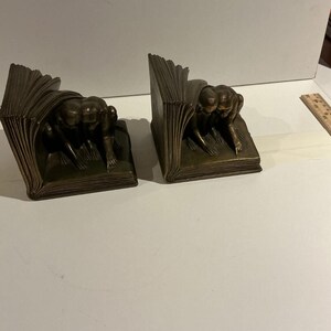 May include: Two bronze bookends, each shaped like an open book with a sculpted figure leaning over the pages. The figures are in a crouching position. The bookends have a dark, antique finish.