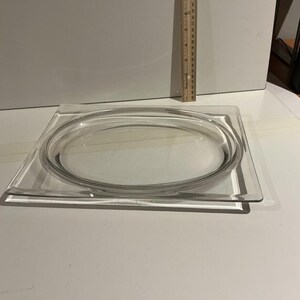 May include: A clear, square glass dish with a rounded, oval-shaped glass insert. A wooden ruler is positioned vertically in the background. The dish is on a white surface. The dish measures approximately 10 inches in length.