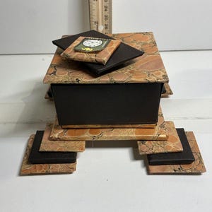 May include: A decorative box with a layered design. The box features a marbled pattern in shades of orange and brown, with black accents. A small square with a clock design is on top. The box is approximately 4 inches wide.