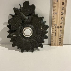 May include: A dark bronze doorbell button featuring a sunflower and bee design. The button has a white centre and is approximately 7.6 cm in diameter. The doorbell is made of metal and has a vintage aesthetic.