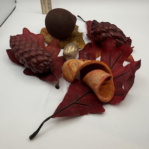May include: A collection of decorative autumn items including two dark brown pinecones, a round dark brown sphere, a metallic silver walnut, and a curled orange wood shaving, all resting on red fabric leaves, with a ruler showing 7.6 cm.