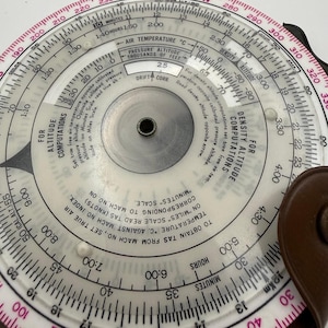 May include: A circular flight computer with a white face, black text, and a red outer ring. It features various scales for altitude, air temperature, and density altitude computations, with a central metal pivot and a brown leather-like case.