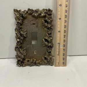 May include: A decorative brass-colored light switch cover with a floral design. The cover has a rectangular shape with a single switch opening and two screw holes. A ruler is next to the cover, showing inches.