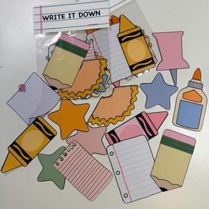 以下が含まれることがあります： 鉛筆、クレヨン、星、メモ帳など、学校用品の形をしたカラフルな紙の切り抜きコレクション。「WRITE IT DOWN」の文字が透明なビニール袋に印刷されています。