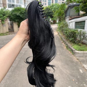 May include: A black hairpiece with a claw clip attachment. The hairpiece is wavy and appears to be synthetic. The claw clip is black and has multiple teeth for securing the hairpiece to the hair.