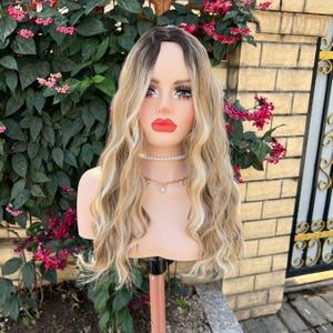 May include: A blonde wig with dark roots and wavy texture is displayed on a mannequin head. The wig has a natural look with varying shades of blonde. The mannequin is wearing a pearl necklace and a gold chain necklace with a pendant.
