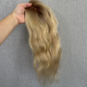 May include: A long, wavy hair topper featuring a rooted design that transitions from a dark brown shade at the base to a bright, multi-tonal blonde throughout the lengths. The hairpiece has a natural-looking part and soft, flowing waves.