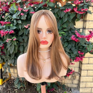 May include: A mannequin head with long, light brown hair and bangs. The mannequin has full makeup, including red lipstick and eye makeup. It is wearing a pearl necklace and a gold chain necklace. The background features green foliage and pink flowers.