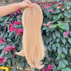 May include: A light blonde hair topper with long, straight strands. The hairpiece is held up against a backdrop of green foliage and vibrant pink flowers, showcasing its color and texture. The hairpiece appears to be made of human hair.