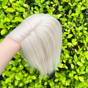 Può includere: Un topper per capelli biondo chiaro con un cuoio capelluto dall'aspetto naturale. I capelli sono lisci e sembrano essere capelli umani. Il topper è tenuto su uno sfondo di fogliame verde.