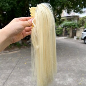 May include: A light blonde hair extension with a claw clip. The hair is straight and appears to be synthetic. The clip is a light beige color and is holding the hair together.