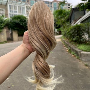 May include: A blonde and light brown hair extension piece with wavy strands. The hair piece is held up against a blurred outdoor background. The hair piece has a mix of light and dark tones.