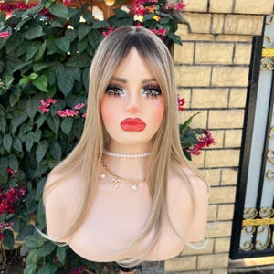 May include: A mannequin head with a long, layered wig in a blend of blonde and dark brown. The mannequin has full makeup, including bold eye makeup and red lipstick. A pearl necklace and a gold chain with charms adorn the neck.