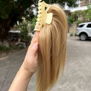 May include: A light blonde hair extension with a claw clip. The hair is straight and appears to be synthetic. The claw clip is a light beige color and is holding the hair extension together.