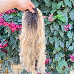 May include: A blonde wig with long, wavy hair. The wig has a dark brown root that fades into a light blonde color. The wig is held up against a background of green leaves and pink flowers.