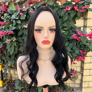May include: A mannequin head with a black wavy wig. The mannequin has full makeup, including red lipstick, and is wearing a pearl necklace and a gold chain. The background features green foliage and pink flowers.