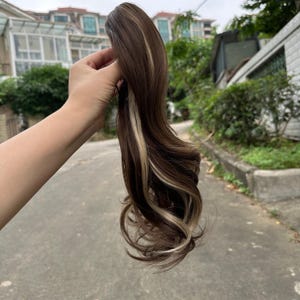 May include: A brown and blonde hair extension with wavy curls. The hairpiece features dark brown strands with streaks of blonde highlights. The hair is held up against a blurred outdoor background.