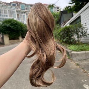 May include: A bundle of wavy, light brown hair extensions with highlights. The hair is held up against a blurred outdoor background. The hair has a natural look with varying shades of brown and blonde.