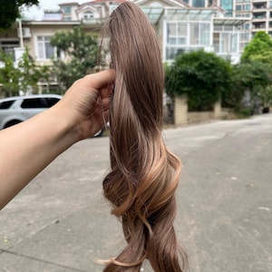May include: A brown and blonde ombre hair extension held up against an outdoor background. The hair is long, wavy, and has a natural look. The colors blend from a dark brown at the top to a lighter blonde at the ends.