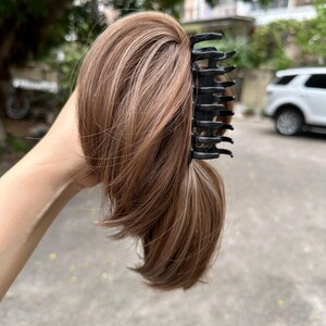 May include: A brown and blonde hairpiece with a black claw clip. The hairpiece is held up, showing the texture and color variations. The clip is open, ready to attach to the hair. The hairpiece appears to be synthetic.