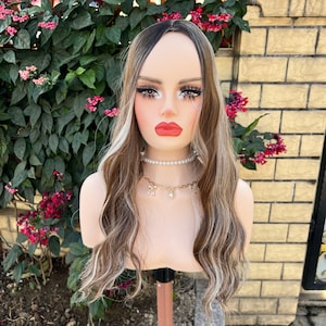 May include: A mannequin head with a long, wavy wig featuring dark roots and blonde highlights. The mannequin has full makeup, including bold red lipstick and long eyelashes. A pearl necklace with gold charms adorns the neck. The background includes greenery and a brick wall.