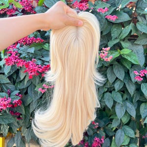 May include: A blonde hair topper with long, layered strands. The hairpiece has a natural-looking part and is held up against a backdrop of green leaves and pink flowers. The hair color is a light, warm blonde.