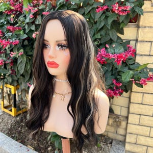 May include: A mannequin head with a long, wavy wig featuring dark brown hair with blonde highlights. The mannequin has full makeup, including red lipstick and a pearl necklace. The wig is styled with loose waves.