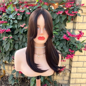 May include: A brown wig with long, straight hair and layered bangs is displayed on a mannequin head. The wig has a dark brown color with a lighter brown highlight. The mannequin is wearing a pearl necklace and a gold chain necklace with charms. The background features green foliage and pink flowers.