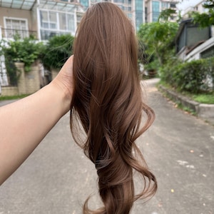 May include: A brown hair extension with wavy ends. The hairpiece is held up against a blurred outdoor background. The hair is a medium brown color and appears to be synthetic.
