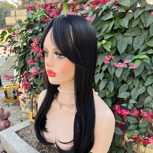 May include: A black wig with long, straight hair and bangs. The wig is styled on a mannequin head with a fair complexion, red lipstick, and a pearl necklace. The background features green foliage and pink flowers.