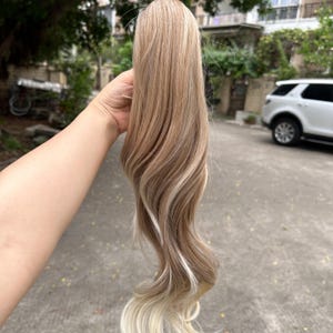 May include: A blonde and light brown hair extension with wavy ends. The hairpiece has highlights and lowlights, creating a natural, multi-dimensional look. The hair is held up by a hand.