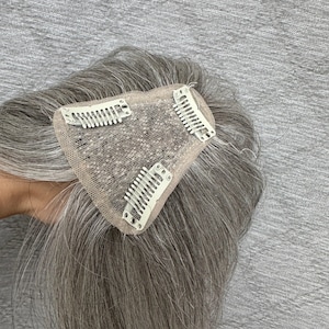 Könnte beinhalten: Ein Haarteil mit glattem, pfeffer-und-salz-grauem Haar, das an einer dreieckigen, beigefarbenen Netzbasis befestigt ist. Die Unterseite der Basis enthält drei metallene Schnappclips zur sicheren Befestigung am vorhandenen Eigenhaar.