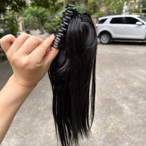 May include: Black hair extension with a claw clip. The hairpiece is a dark, solid black color. The claw clip is made of black plastic and is designed to hold the hair extension securely in place. The hair extension is long and straight.