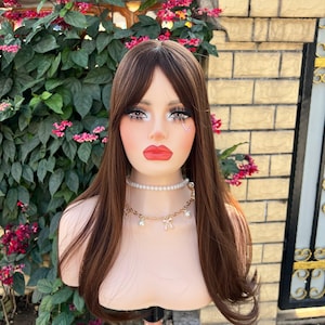 May include: A mannequin head with long, straight, brown hair and bangs. The mannequin is wearing a pearl necklace and a gold chain necklace with bows and pearls. The mannequin has full makeup, including red lipstick and long eyelashes.