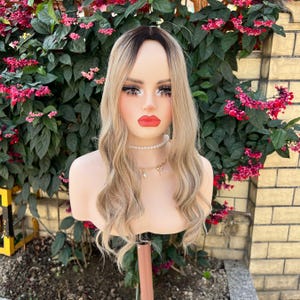 May include: A blonde wig with dark roots and wavy layers is displayed on a mannequin head. The mannequin has full makeup, including red lipstick and long eyelashes. A pearl necklace and gold pendant are visible. The background features pink flowers and a brick wall.