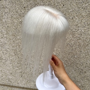 以下が含まれることがあります： 中央に分け目がある、真っ直ぐで鮮やかな白い髪のヘアトッパー。髪は滑らかな質感で、頭頂部から真っ直ぐに垂れ下がっています。このヘアピースは、展示用に白いプラスチック製のスタンドに取り付けられています。