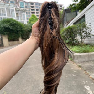 May include: A brown and blonde hair extension with a black claw clip. The hairpiece features a mix of dark brown and light brown highlights, creating a natural look. The clip is designed to securely attach the extension to the hair.