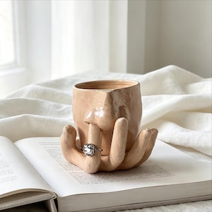 May include: A light peach-coloured ceramic mug shaped like a human face with a nose protruding from the centre. The base of the mug is formed by a sculpted hand with fingers wrapped around the sides. A silver-toned ring is worn on one finger.