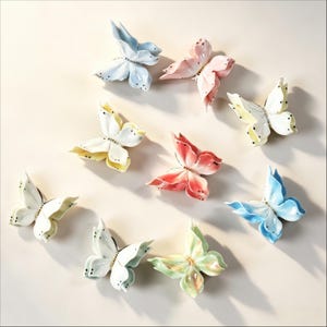 Op de afbeelding: Een verzameling van negen keramische vlinderwanddecoraties in diverse pastelkleuren, waaronder lichtblauw, zachtroze, geel, wit, koraal en lichtgroen. Elke vlinder heeft verfijnde, gebeeldhouwde vleugels met kleine zwarte stippen als accent.