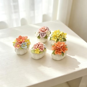 May include: Five small, white ceramic containers, each shaped like a rounded pumpkin, are topped with sculpted, multi-colored floral arrangements. The flowers feature shades of pink, orange, yellow, and blue, accented with green leaves.
