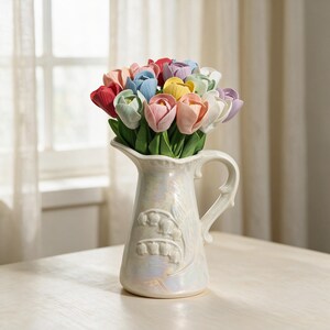 Pode incluir: Um buquê de tulipas artificiais multicoloridas em tons de vermelho, rosa, amarelo, azul e branco com folhas verdes, arranjado em um jarro de cerâmica branco perolado com um padrão floral de lírios do vale em relevo e uma alça.
