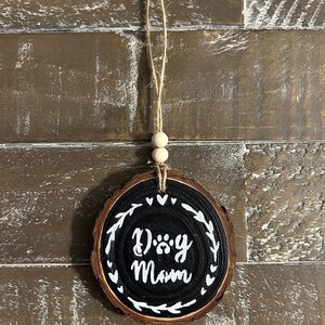 May include: A round, wooden ornament with the words "Dog Mom" in white script on a black background. The ornament is trimmed with a white leaf design and has a twine hanger with two wooden beads. The background is a rustic, wooden surface.