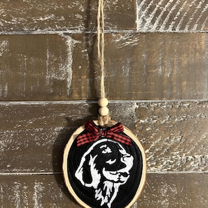 May include: A wooden dog ornament with a black background and a white dog silhouette. The ornament is adorned with a red and black plaid bow and a twine hanger with wooden beads. A rustic, country-style decoration.