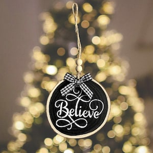 May include: Round black and white Christmas ornament with the word "Believe" in white script. The ornament has a wooden frame, a black and white checkered bow, and a twine hanger with wooden beads. The background is a blurred Christmas tree.
