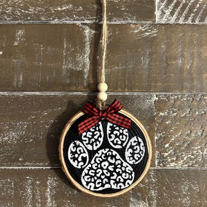 May include: A round, wooden ornament with a black background featuring a white paw print design. A red and black plaid bow is at the top, with a twine loop for hanging. Two small wooden beads are also attached.
