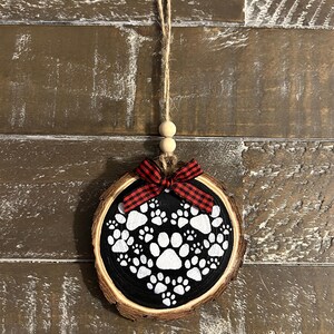 May include: A round, wooden ornament with a black center featuring white paw prints in a heart shape. A red and black plaid bow is at the top, with a jute string for hanging. The ornament is approximately 3 inches in diameter.